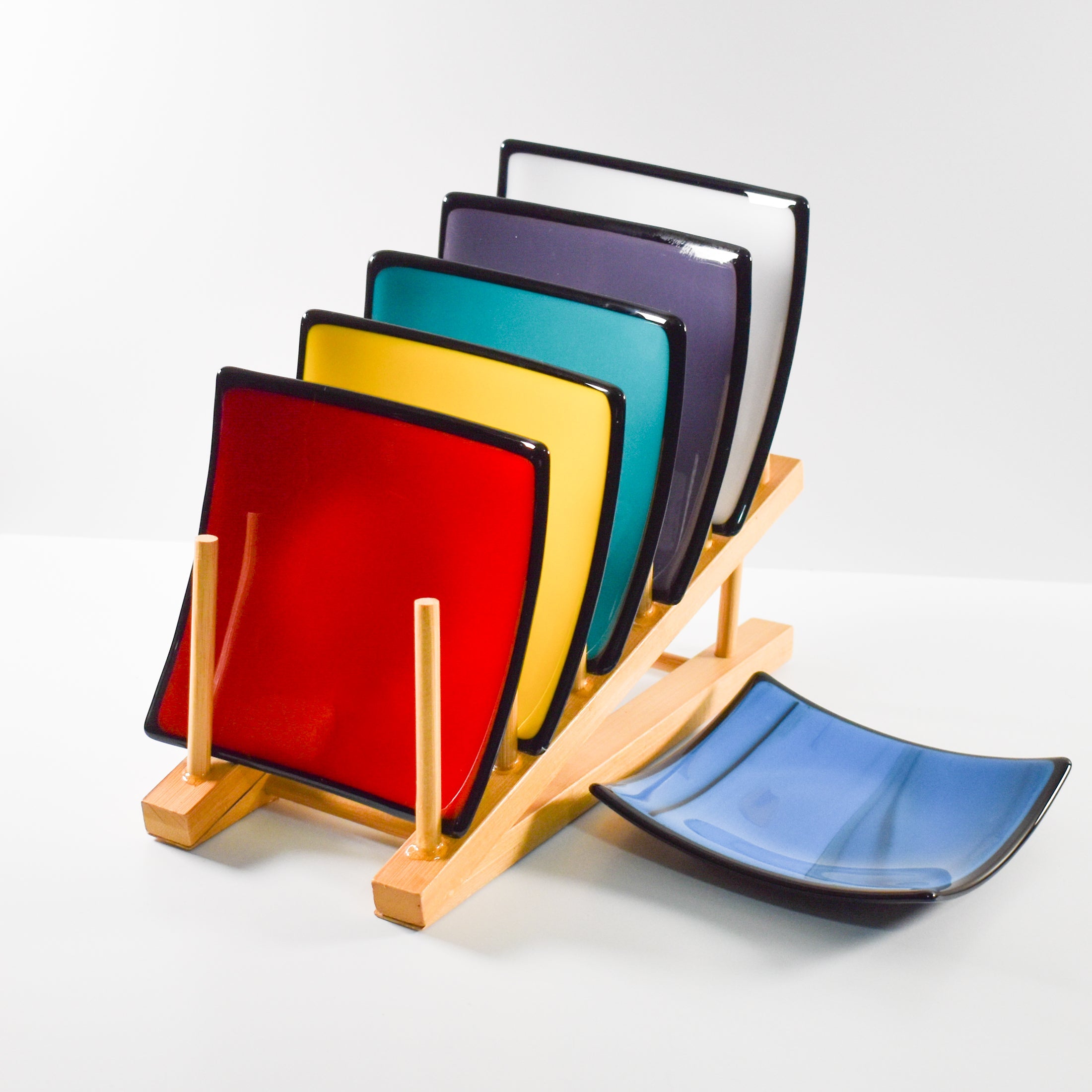 Colorful fused glass dishes in red, yellow, teal, purple, white and blue with black trim, arranged in a wooden holder with one dish in front.