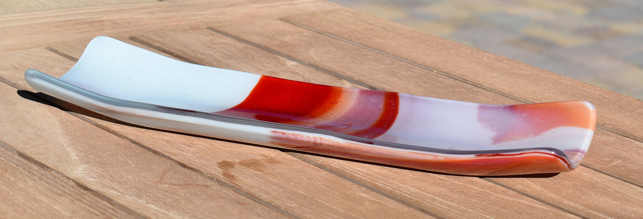 Fused glass tray in long, narrow channel shape made of white, red and orange glass with modern abstract design, resting on wooden slatted table.