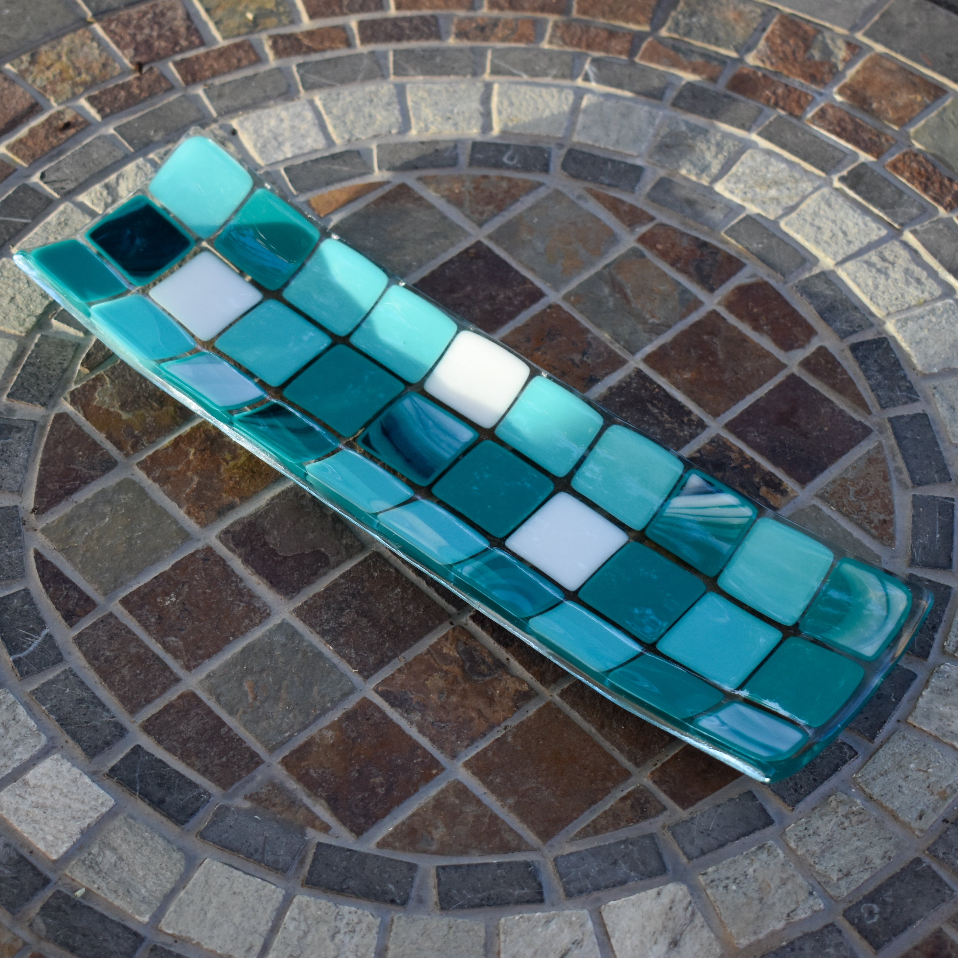 Handmade fused glass tray with blue, teal and white square mosaic pattern, shaped into a long curved dish on a stone tabletop.