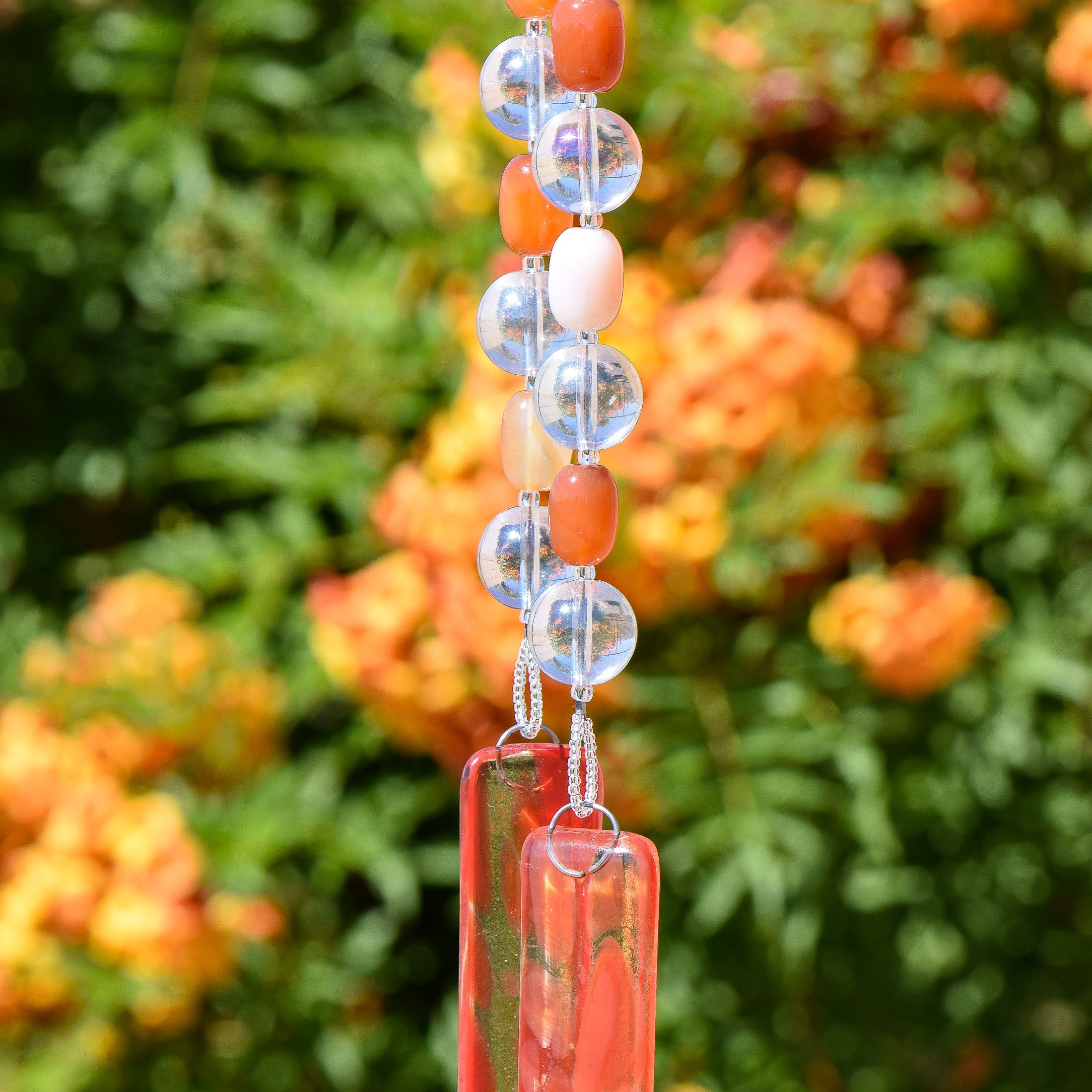 Orange Carnelian agate stone beads paired with large, solid glass clear beads, strung and hanging vertically, anchored by two pieces of orange and clear fused glass. Background is a shrub with orange flowers.