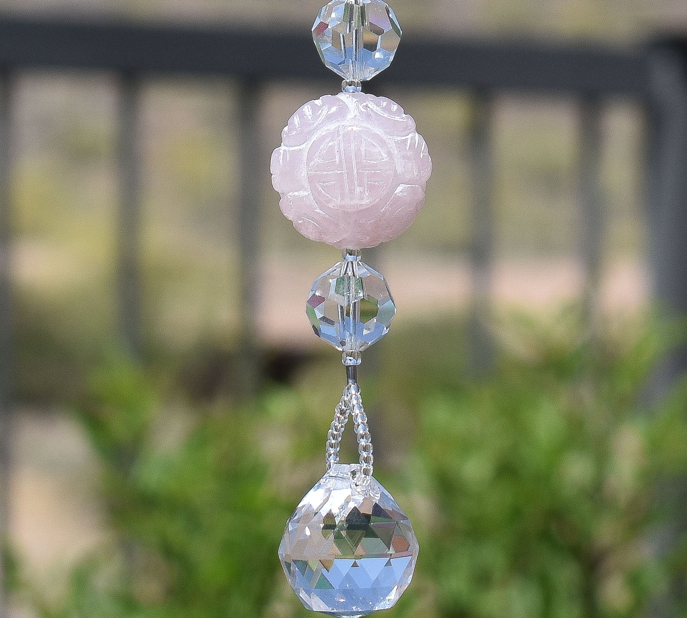 Large round carved rose quartz stone bead flanked by two faceted crystal beads and anchored by a small crystal prism, hanging vertically.