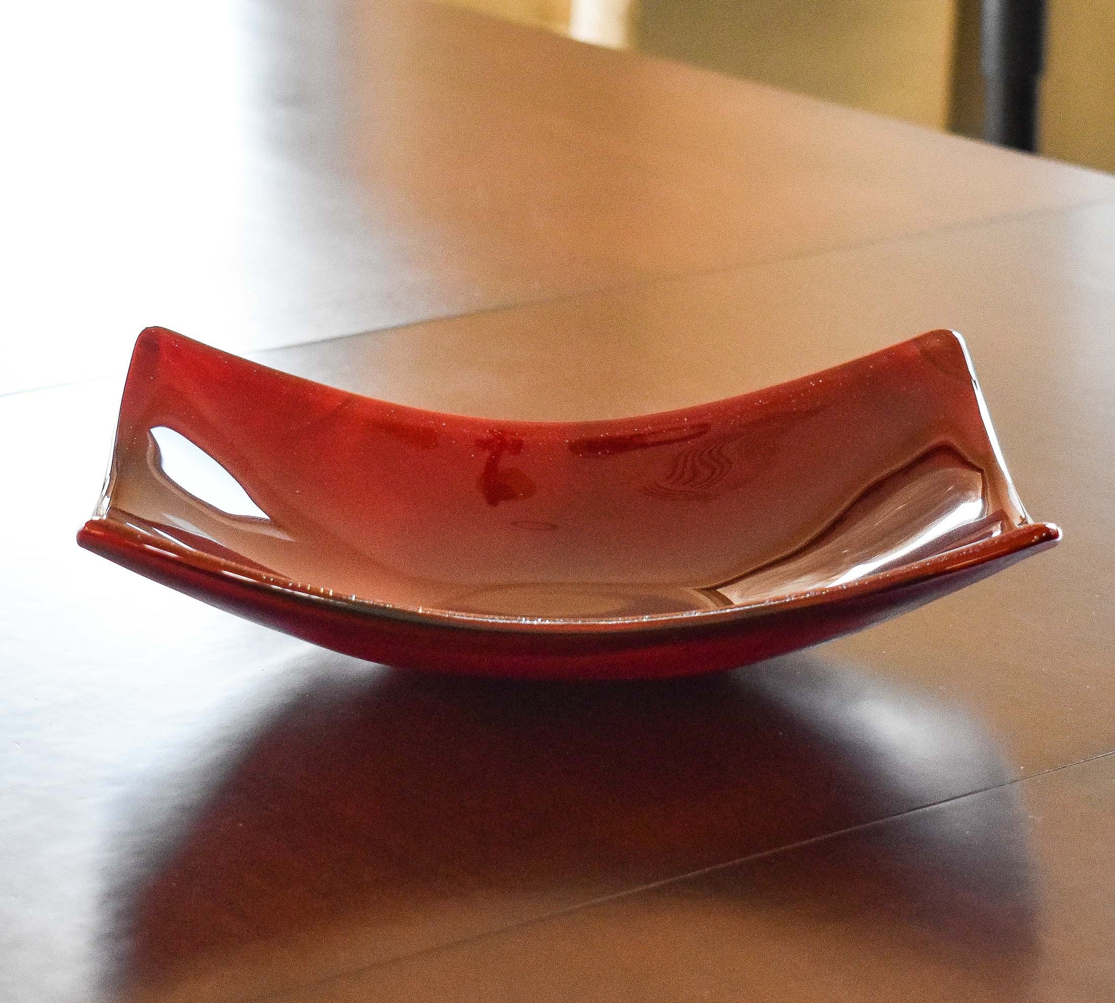 Boat-shaped fused glass dish made of rose red glass, resting on wood table.