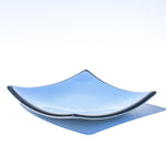 Denim blue square glass dish with black trim and slightly sloped corners, viewed from the side in front of a white background.