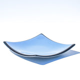 Denim blue square glass dish with black trim and slightly sloped corners, viewed from the side in front of a white background.