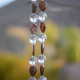 Closer view of two strings of wire covered in Rhyolite stone beads and large clear glass beads hanging vertically against foliage background. The Rhyolite stone beads are tear drop shaped and there is a larger, clear, reflective glass bead in between each stone bead. The stone beads are marked and in shades of brown, tan and gray.