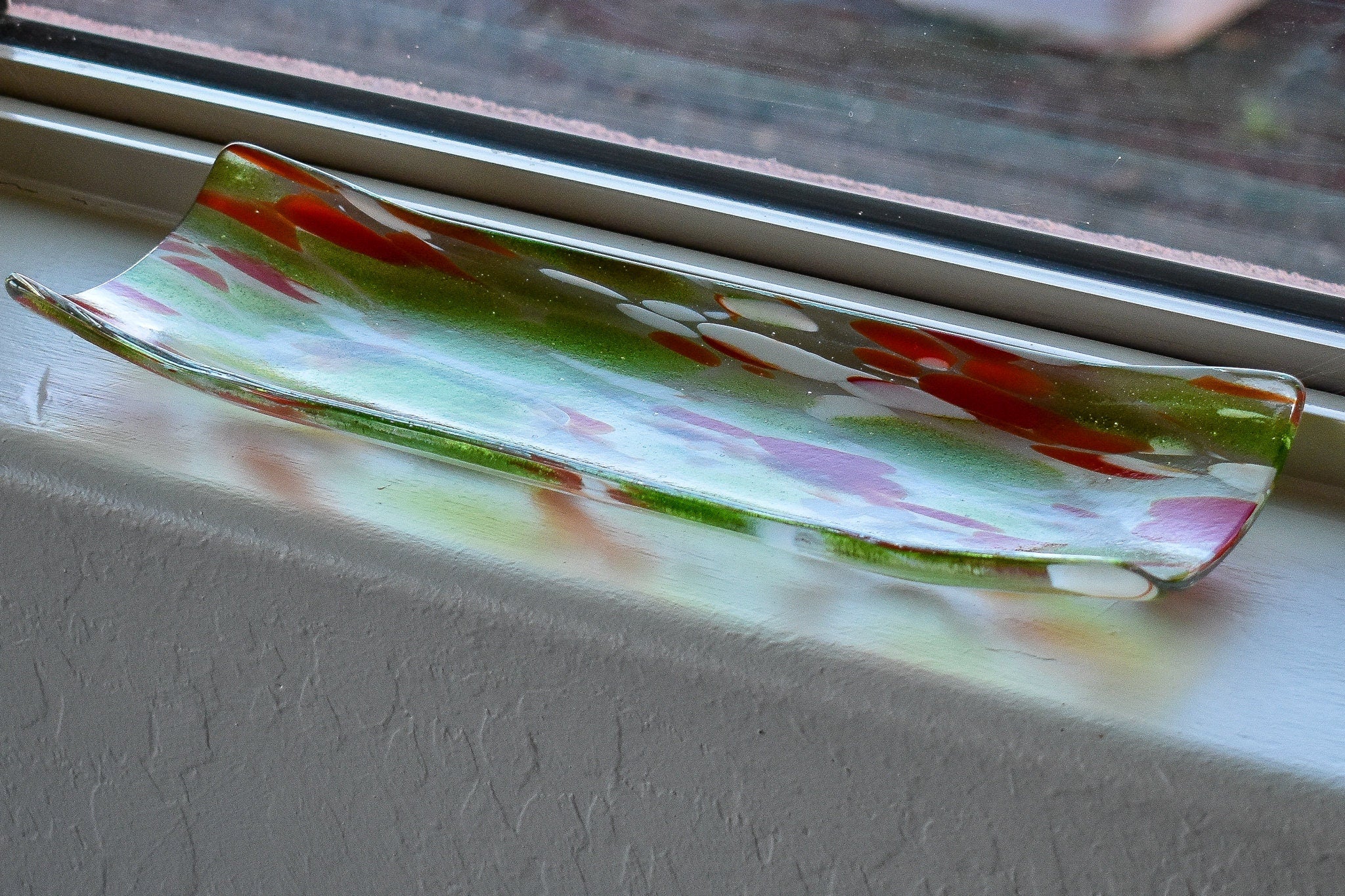 A rectangular glass dish with curved edges. The dish is clear with a blend of colors, including green, red, and white, creating a speckled pattern. It sits on a white window sill.