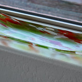 A rectangular glass dish with curved edges. The dish is clear with a blend of colors, including green, red, and white, creating a speckled pattern. It sits on a white window sill.