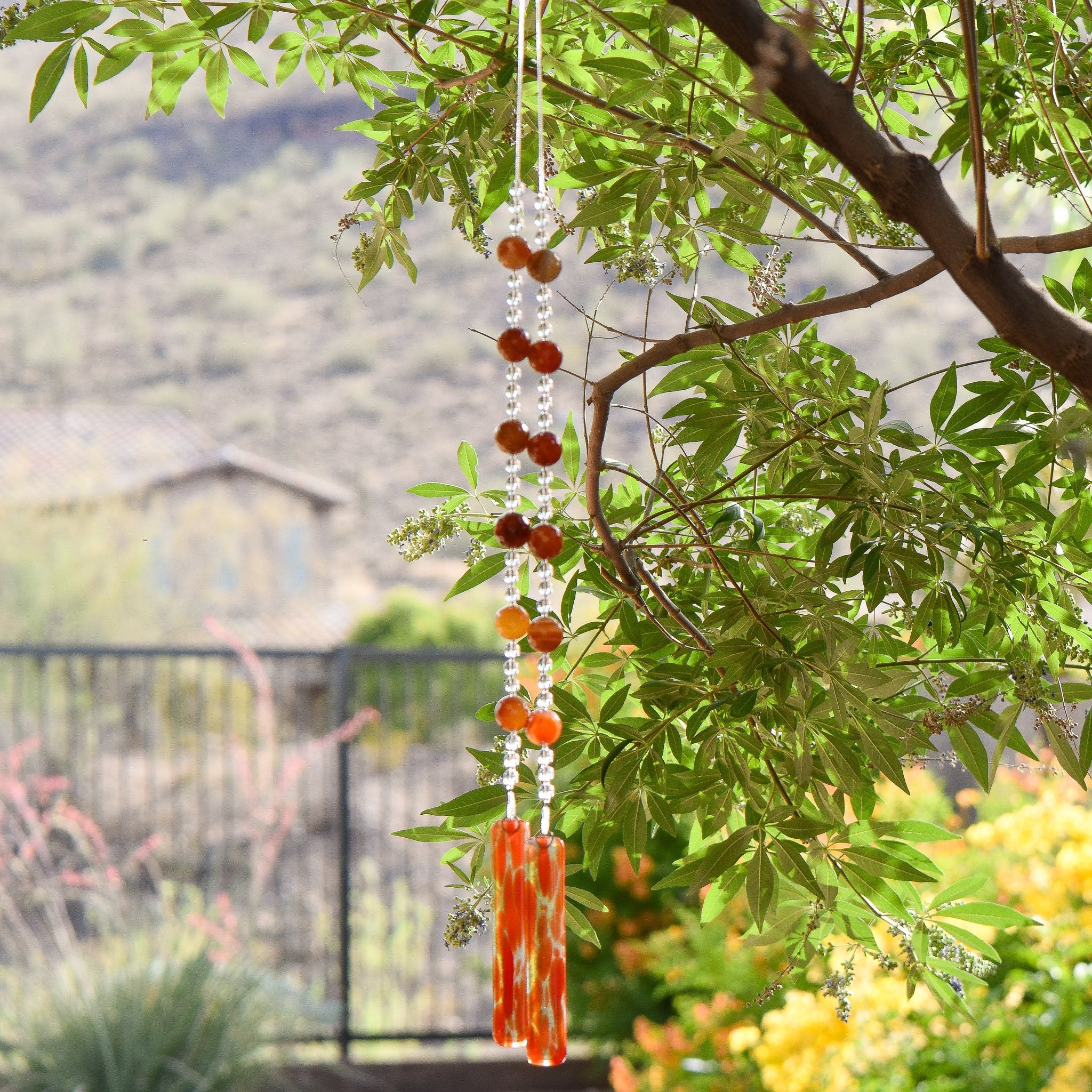 Large, round, faceted carnelian agate stone beads paired with small clear glass beads are hanging vertically from a tree, anchored by two pieces of orange/red/clear glass