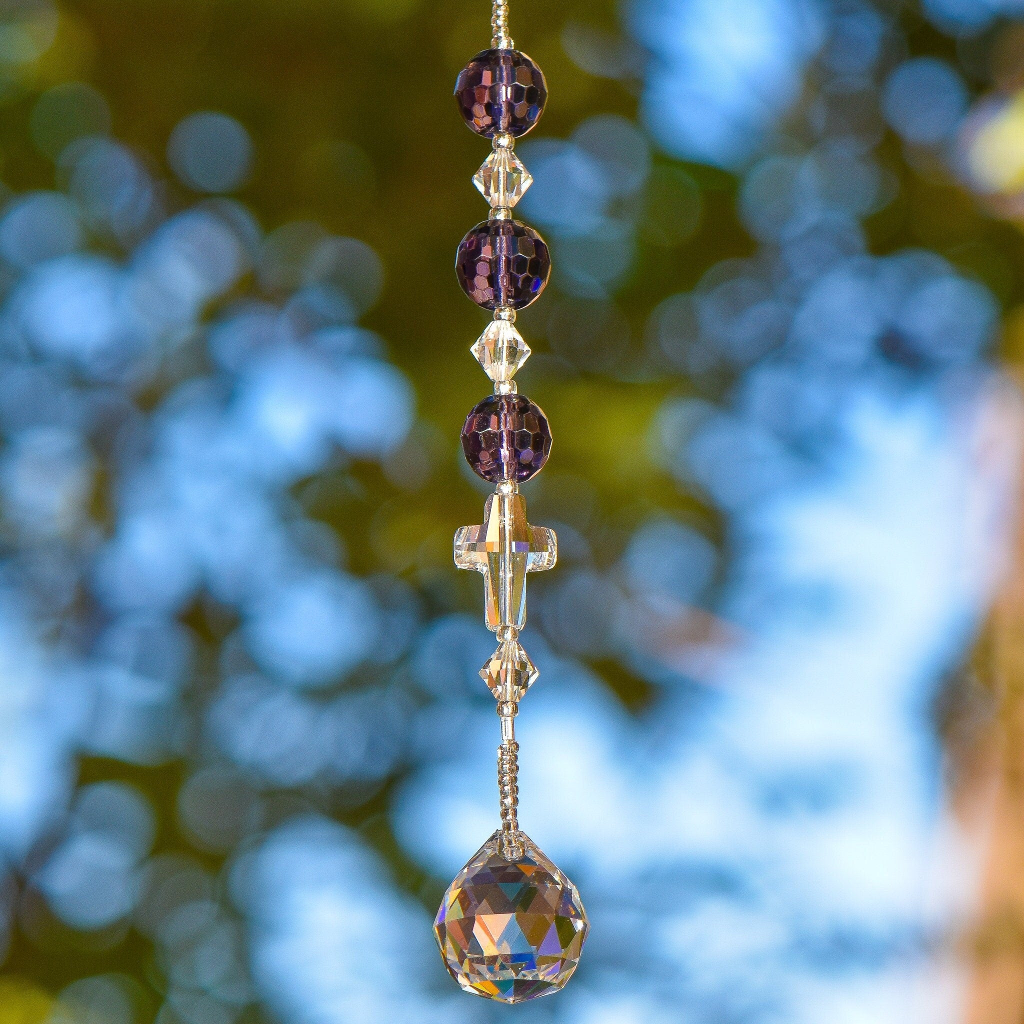 Small suncatcher car charm made with 3 faceted purple glass beads, 3 bicone glass crystal beads and a glass crystal cross, all anchored by a small round prism, hanging vertically from tree.