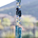 Slices of dark blue agate stone strung with clear and dark aqua glass beads, anchored by two pieces of blue and clear fused glass, hanging from tree.