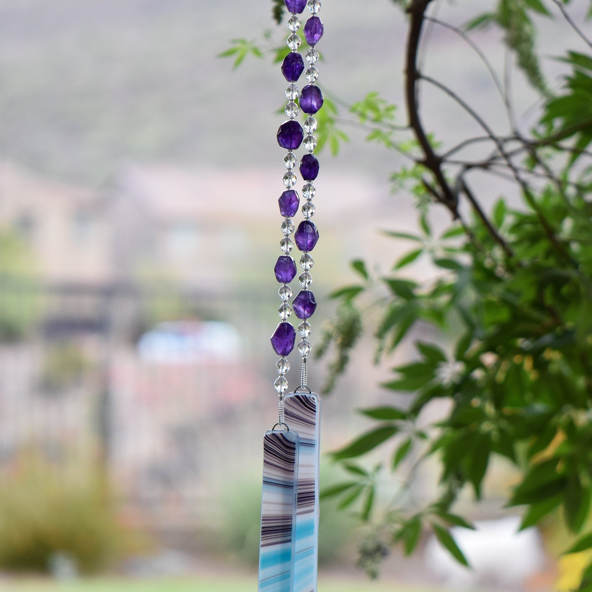 Amethyst stones strung with round crystal beads and anchored by two pieces of blue, white and purple kiln-formed glass, hanging vertically from tree.