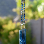 Glass beads, in dark aqua and clear silver, hanging vertically from a tree, anchored by two pieces of blue swirl fused glass.