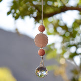 Large round carved rose quartz bead flanked by two smaller pink stone beads and anchored by round crystal prism, hanging from tree.