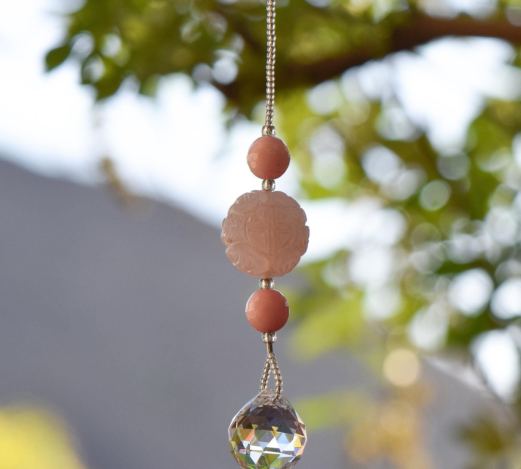 Large round carved rose quartz bead flanked by two smaller pink stone beads and anchored by round crystal prism, hanging from tree.