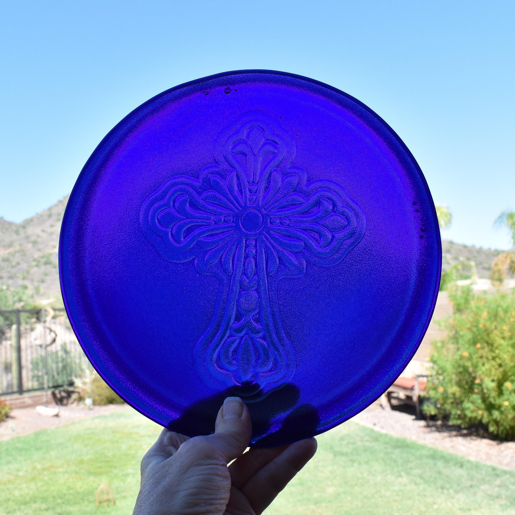 A round, deep blue glass plate with an embossed cross design in the center. The cross has ornate detailing. The plate is held up against a bright blue sky and green grass background.