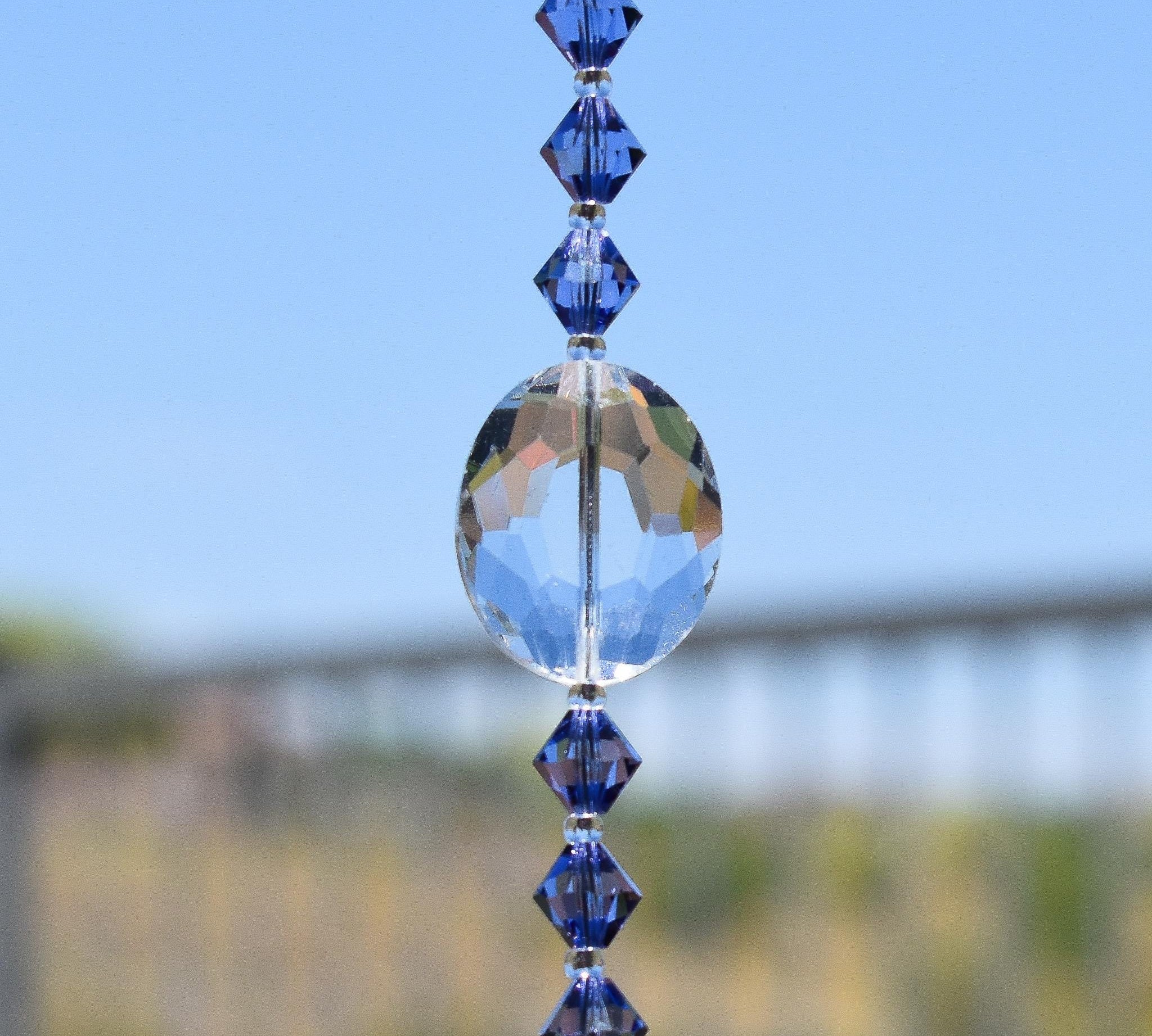 Four small purple crystal bicones in &quot;tanzanite&quot; color flank a large glass crystal oval, strung together and having vertically against blurred fenced background.