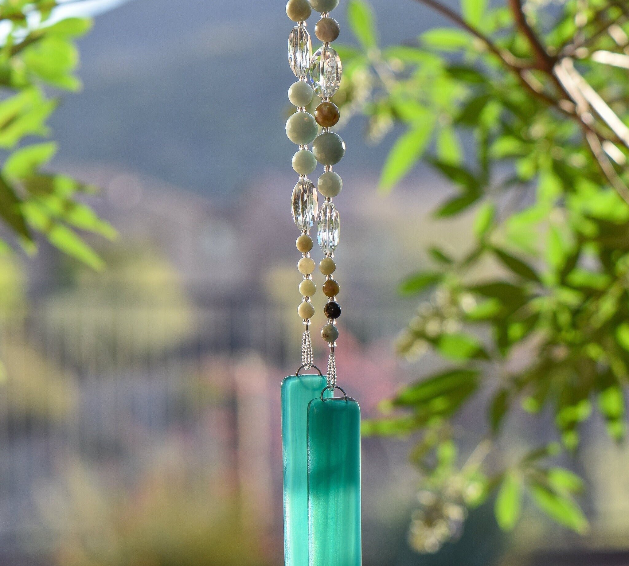 Round amazonite beads, in various sizes, are strung with oval clear crystal beads and hanging vertically from a tree, anchored by two pieces of teal green glass