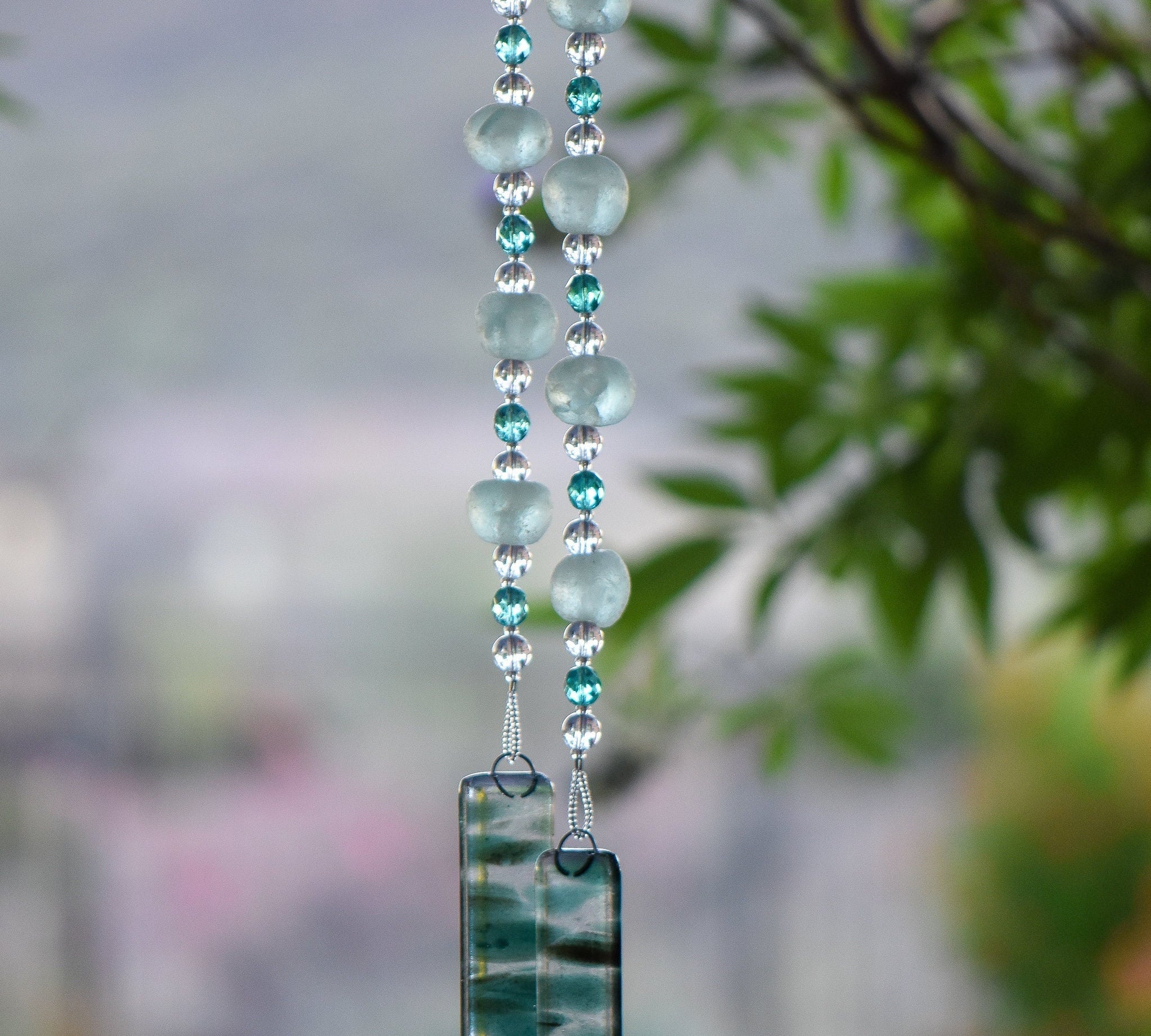 Large sea glass beads from Ghana are strung with smaller clear and aqua-colored glass beads, hanging vertically from tree, anchored by two pieces of green and clear fused glass.