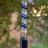 Large round blue lapis lazuli beads strung with crystal ovals, anchored by two pieces of navy blue fused glass, hanging from tree.