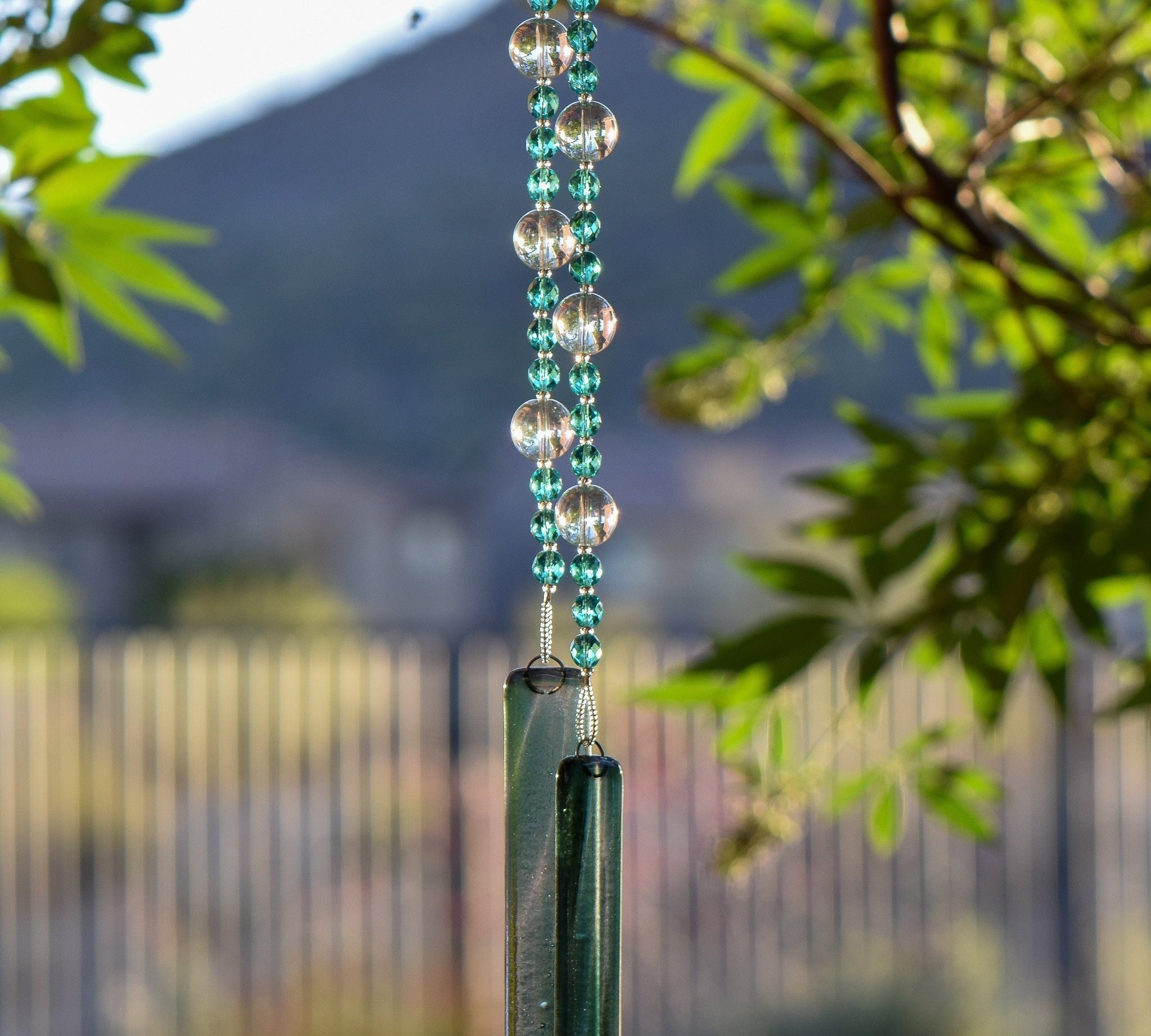 Small, faceted aqua green glass beads strung with larger solid clear glass beads, hanging vertically from a tree, anchored by two pieces of green and clear fused glass.