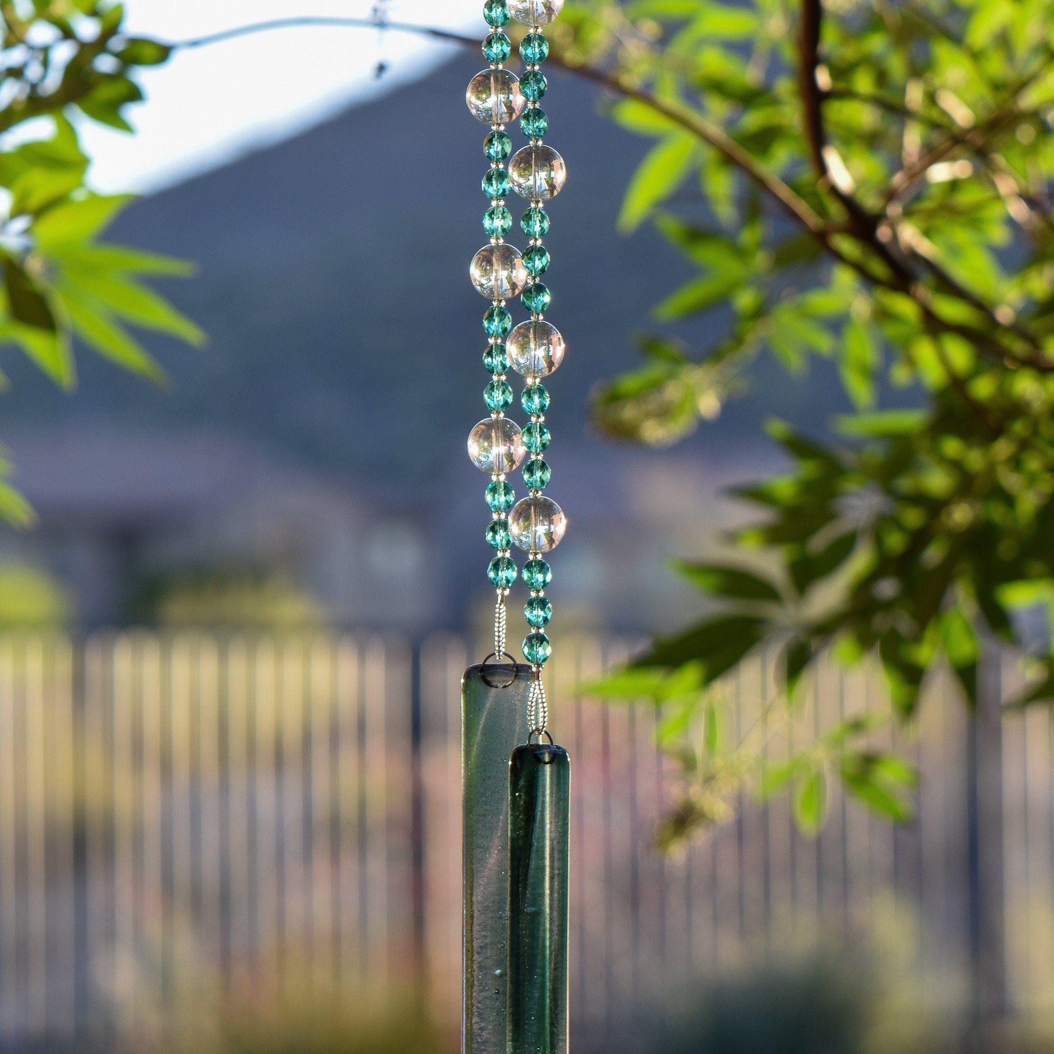 Small, faceted aqua green glass beads strung with larger solid clear glass beads, hanging vertically from a tree, anchored by two pieces of green and clear fused glass.