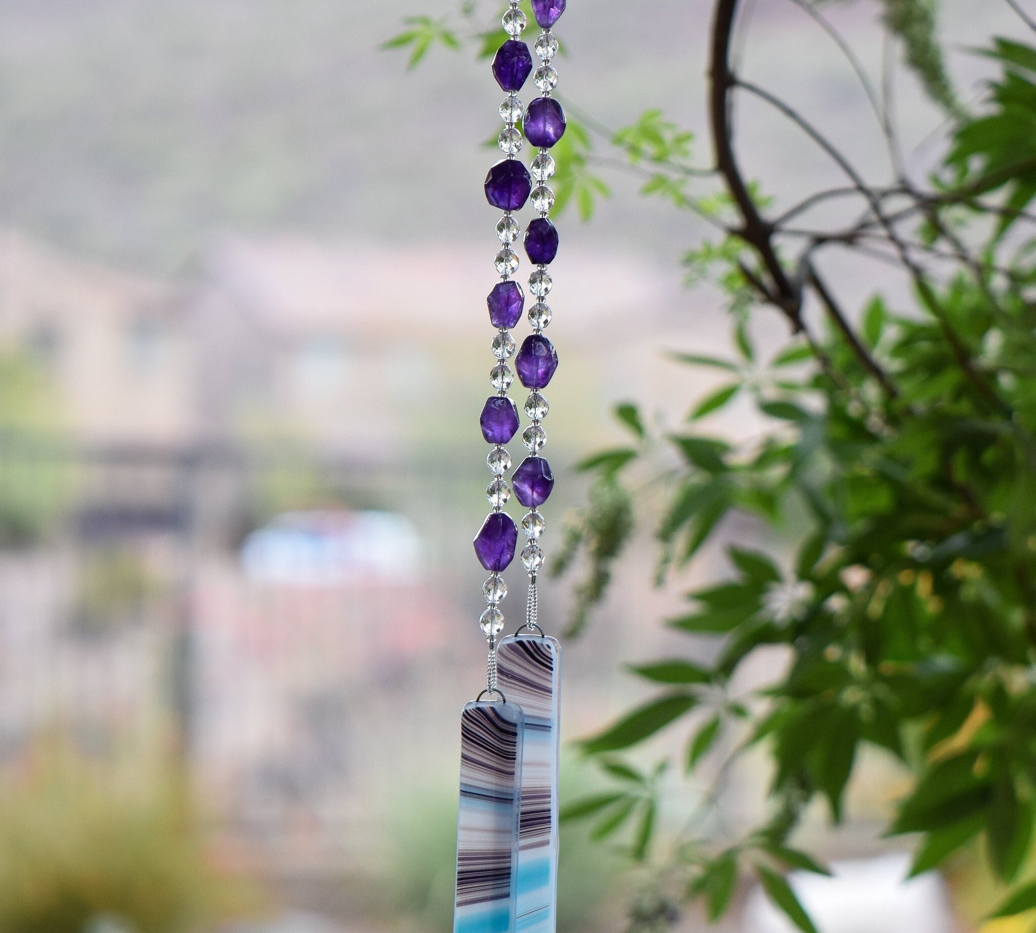 Amethyst stones strung with round crystal beads and anchored by two pieces of blue, white and purple kiln-formed glass, hanging vertically from tree.