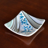 6 inch square fused glass dish with swirl pattern and small flecks of white and blue glass, sitting on wood table