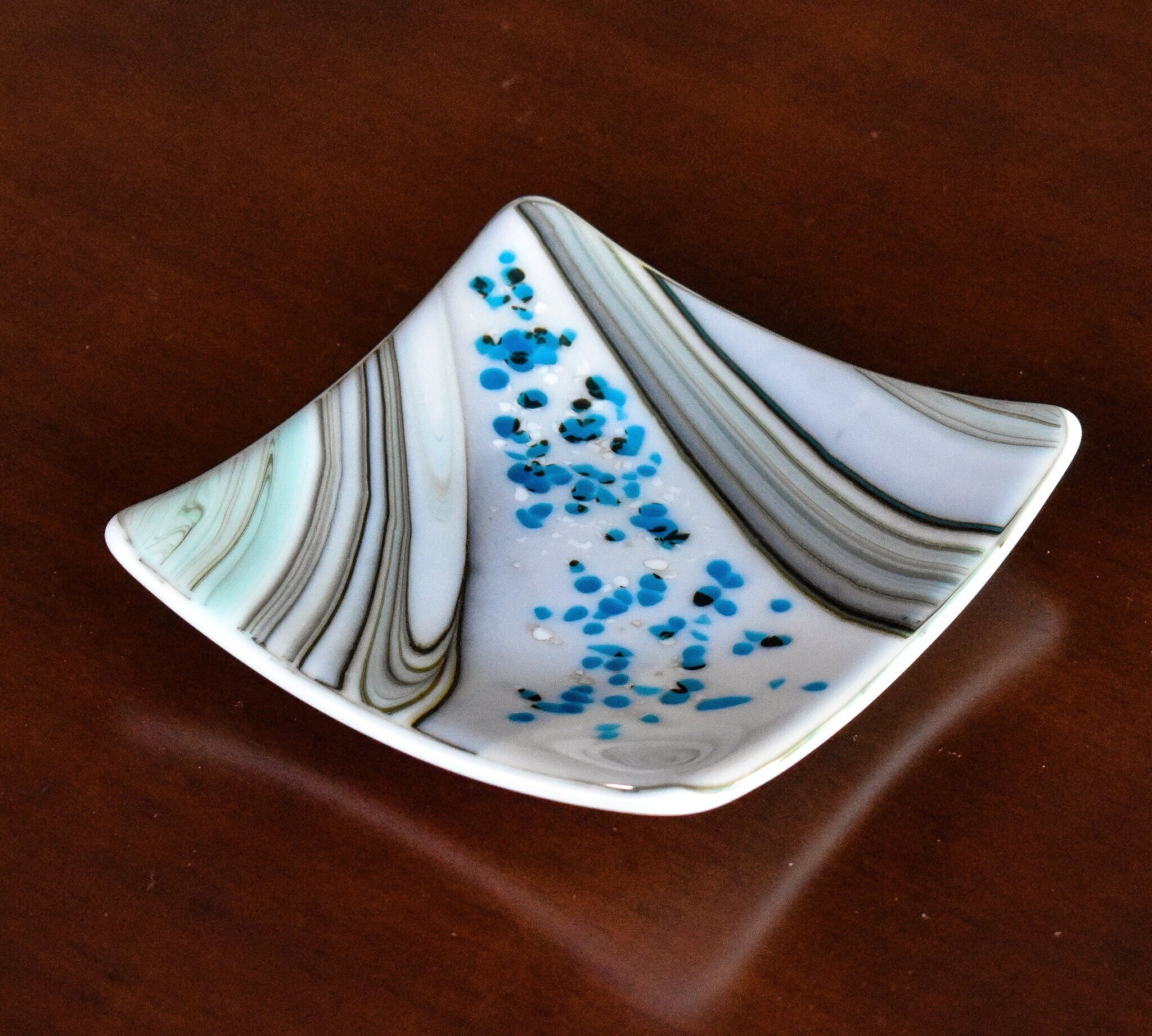 6 inch square fused glass dish with swirl pattern and small flecks of white and blue glass, sitting on wood table