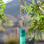 Round amazonite beads, in various sizes, are strung with oval clear crystal beads and hanging vertically from a tree, anchored by two pieces of teal green glass