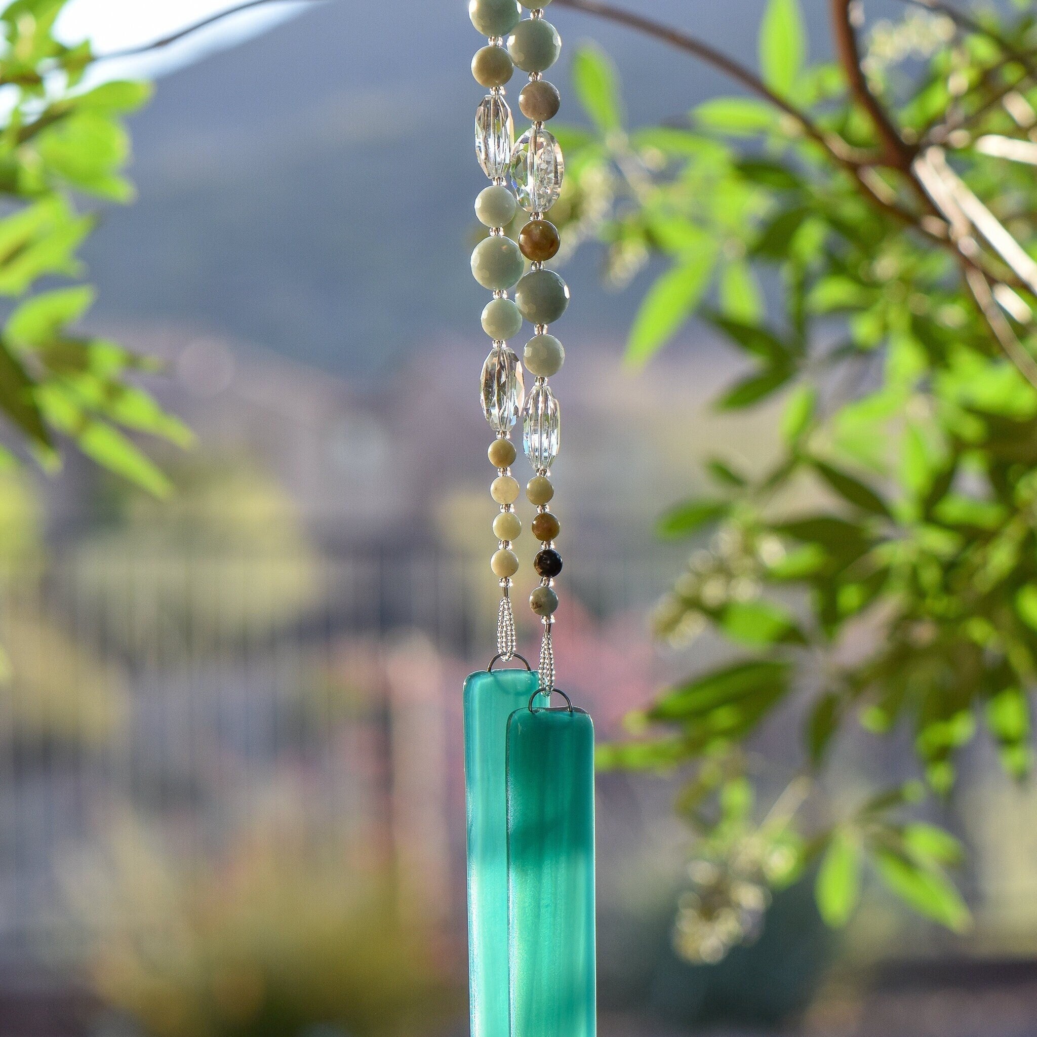 Round amazonite beads, in various sizes, are strung with oval clear crystal beads and hanging vertically from a tree, anchored by two pieces of teal green glass