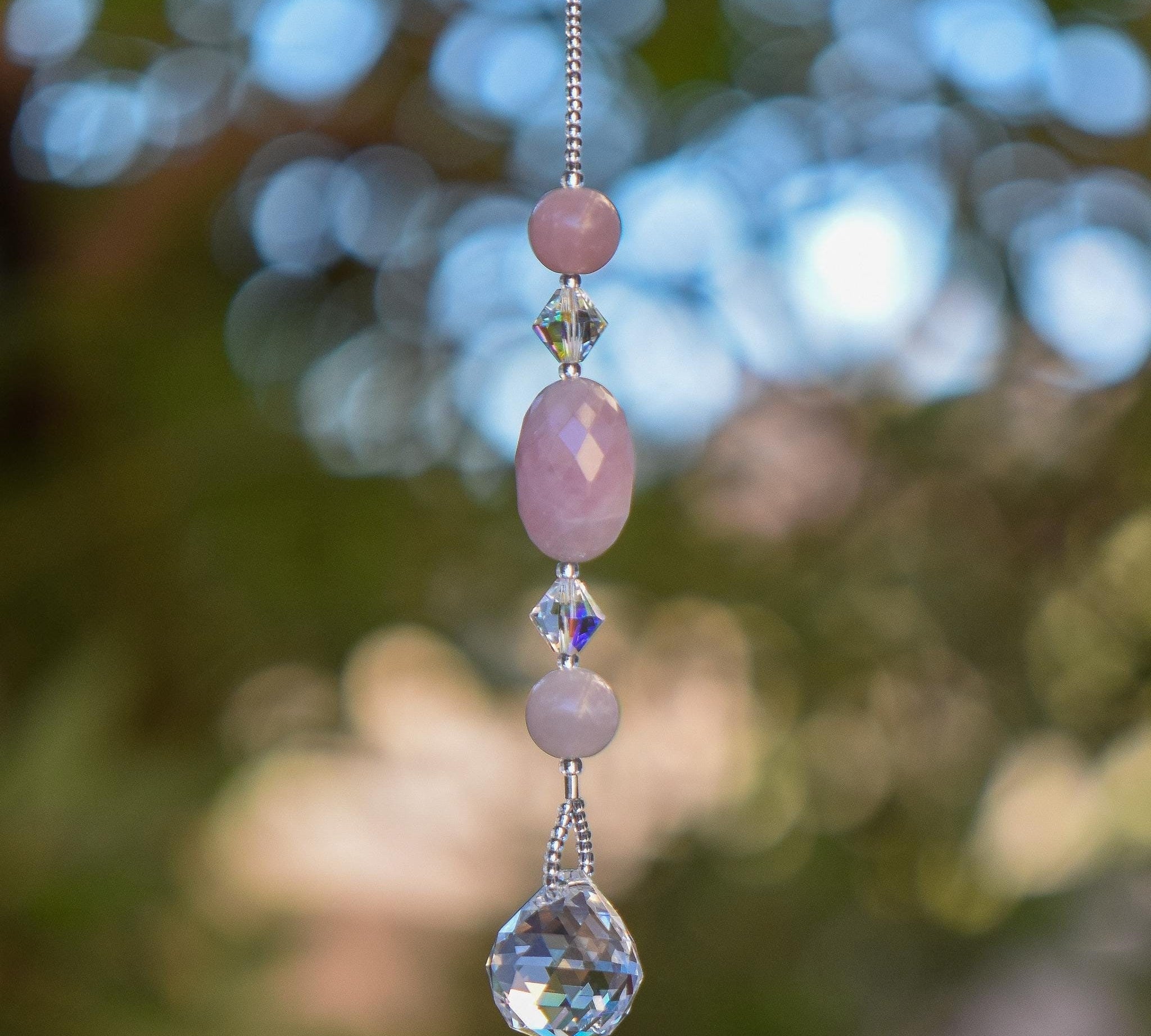 Large oval rose quartz bead flanked by Austrian crystal and two smaller round rose quartz beads, hanging vertically from a tree, anchored by round crystal prism