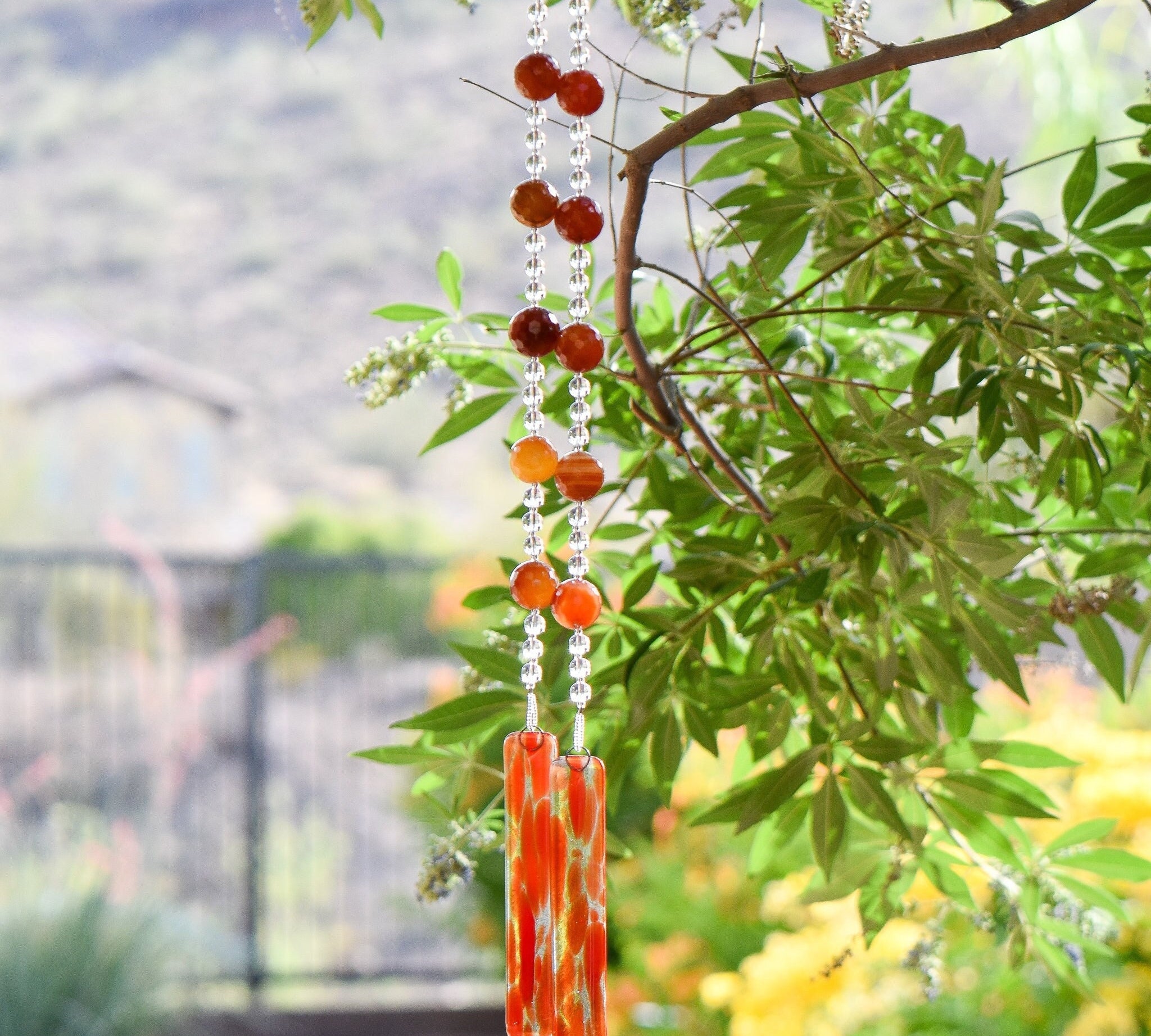 Large, round, faceted orange/red carnelian agate stones strung with glass beads, hanging from a tree, anchored by two pieces of orange, red and clear fused glass