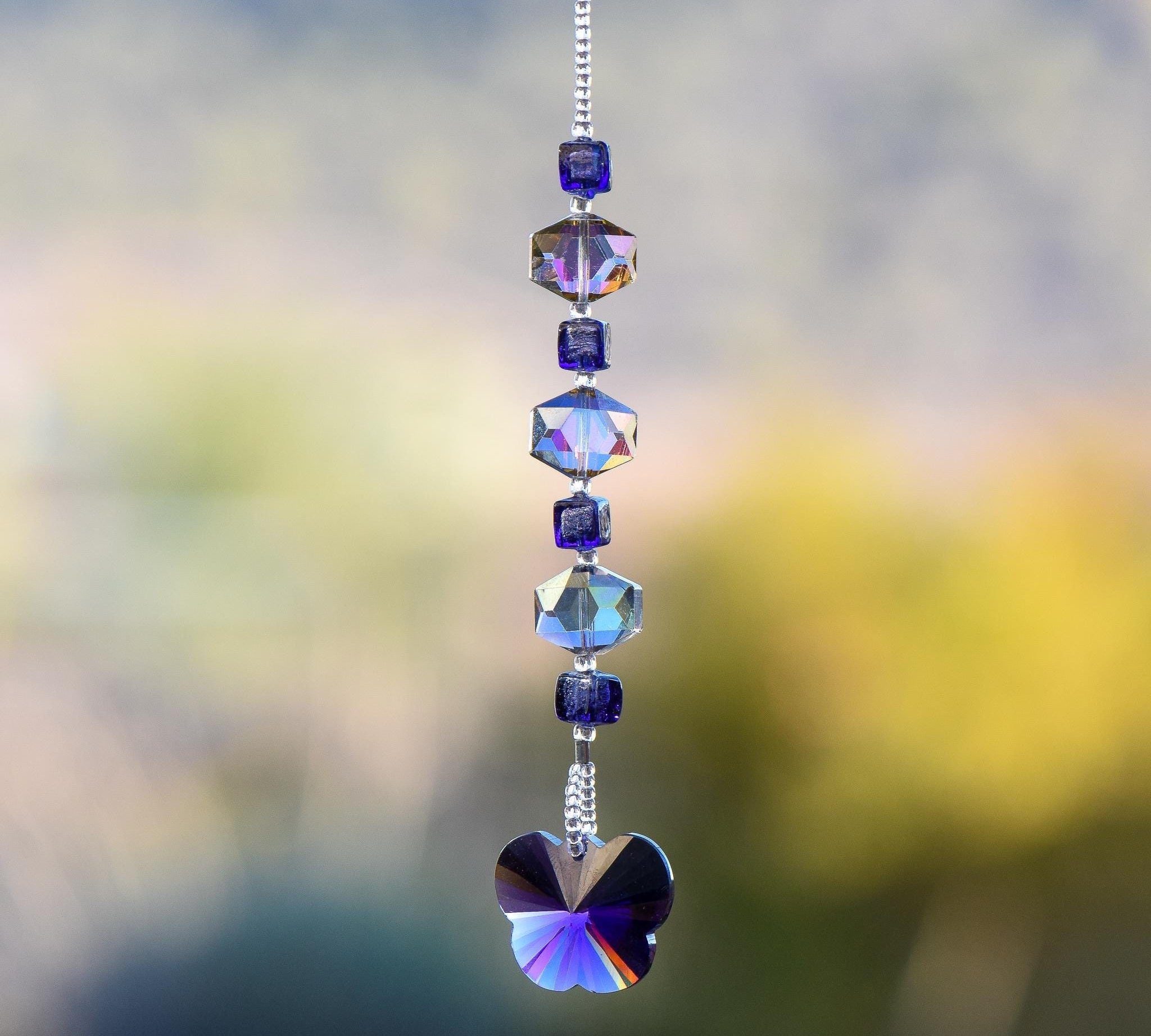 A handmade sun catcher featuring purple and iridescent crystal beads, finished with a faceted crystal butterfly pendant that reflects light in purple, blue, and gold tones. Suspended from a silver-tone chain against a blurred outdoor background.