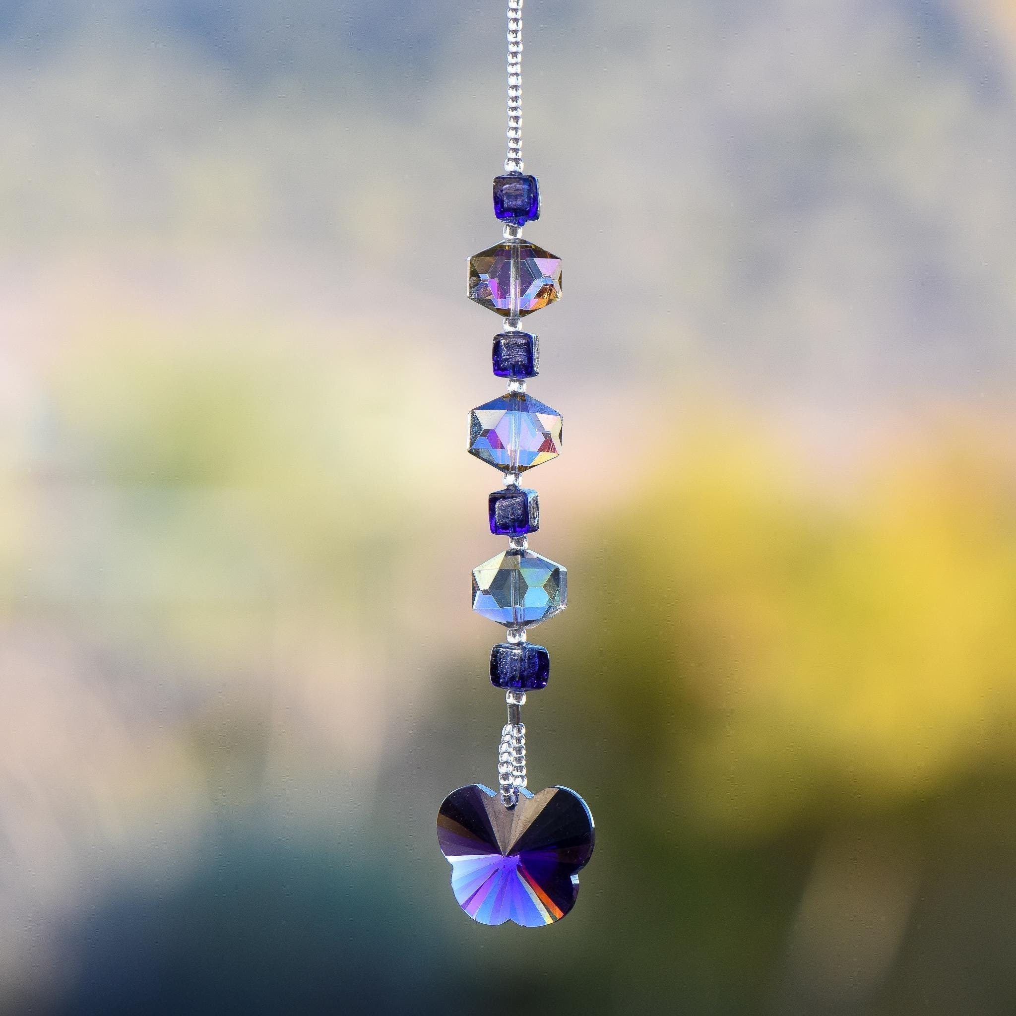 A handmade sun catcher featuring purple and iridescent crystal beads, finished with a faceted crystal butterfly pendant that reflects light in purple, blue, and gold tones. Suspended from a silver-tone chain against a blurred outdoor background.