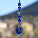 Handmade sun catcher featuring cobalt blue murano glass beads, faceted iridescent crystal cross and a blue/metallic glass disc, hanging vertically against blurred background