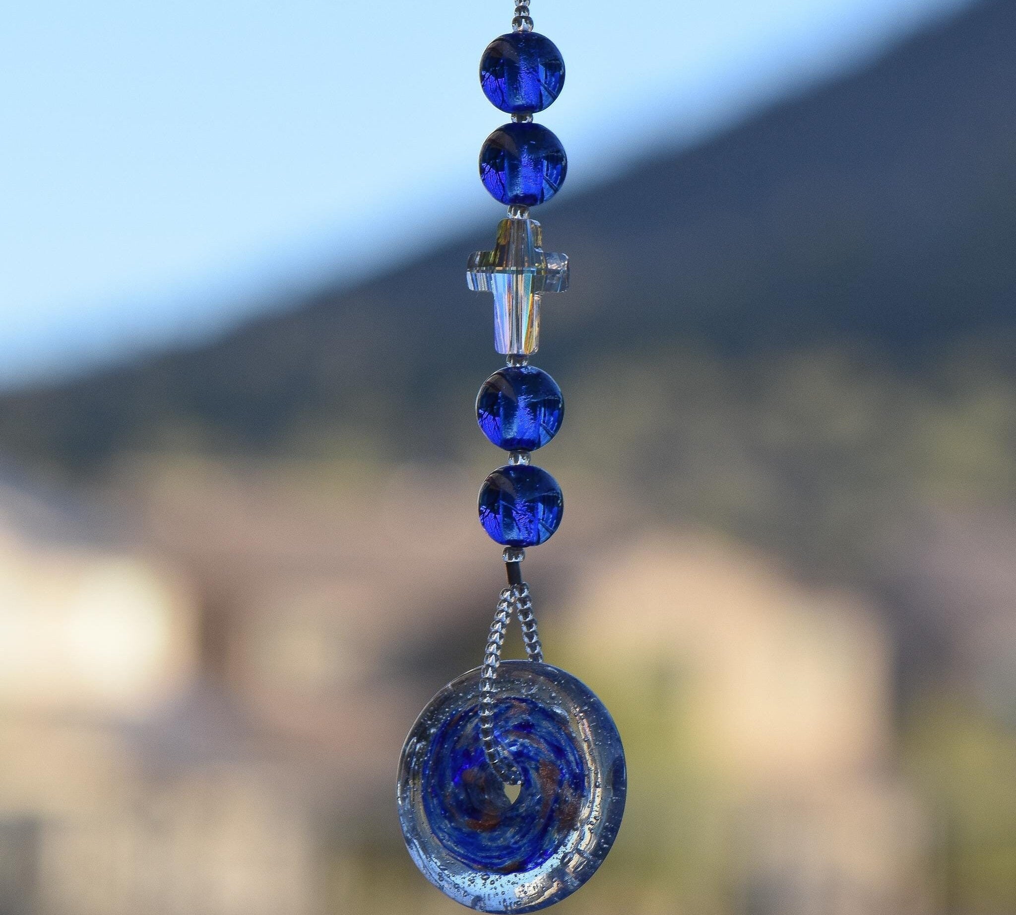 Handmade sun catcher featuring cobalt blue murano glass beads, faceted iridescent crystal cross and a blue/metallic glass disc, hanging vertically against blurred background