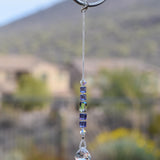 Small sun catcher hanging vertically against blurred background, made with purple crystal cube beads, a large emerald cut crystal and a round crystal prism anchor. The top of the sun catcher is hanging from a hook.