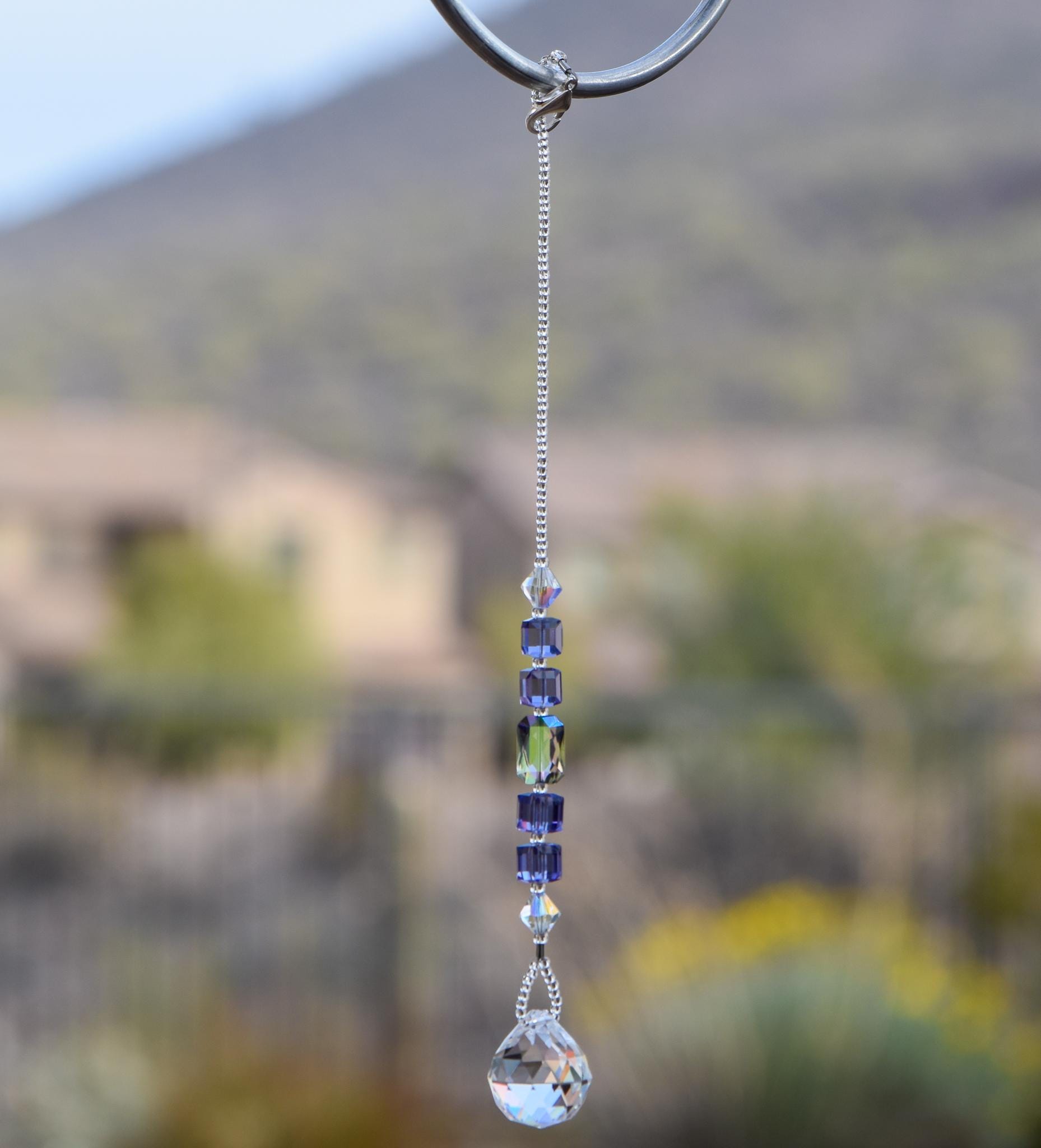 Small sun catcher hanging vertically against blurred background, made with purple crystal cube beads, a large emerald cut crystal and a round crystal prism anchor. The top of the sun catcher is hanging from a hook.
