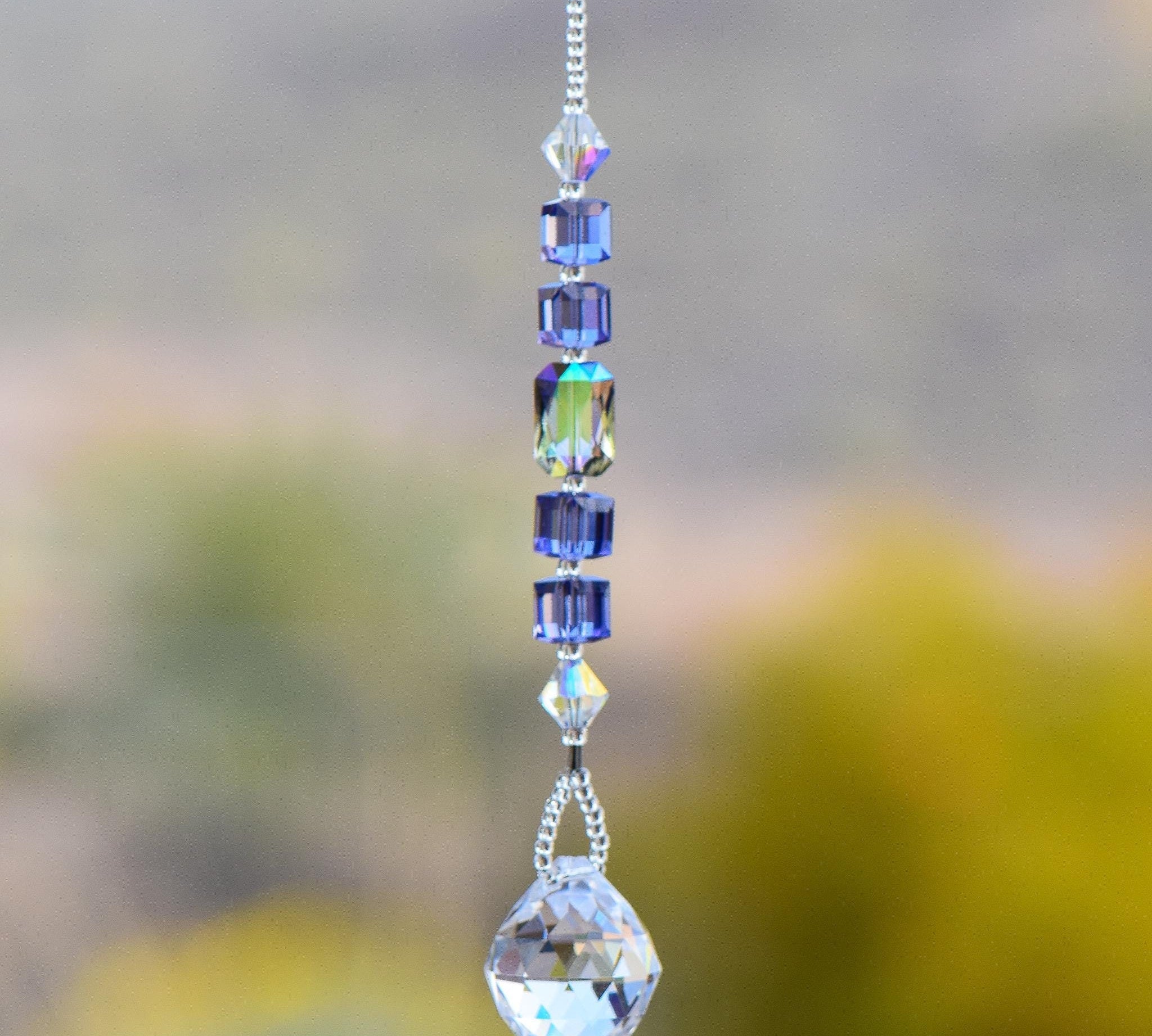 Small sun catcher hanging vertically against blurred background, made with purple crystal cube beads, a large emerald cut crystal and a round crystal prism anchor.