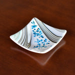 Six inch square glass tray with modern design on wood table.Tray is white with swirls and flakes of aqua, cream, tan and gray.