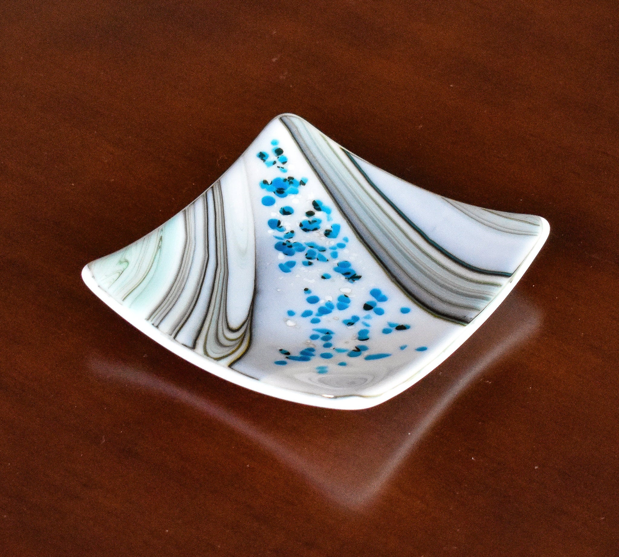 Six inch square glass tray with modern design on wood table.Tray is white with swirls and flakes of aqua, cream, tan and gray.