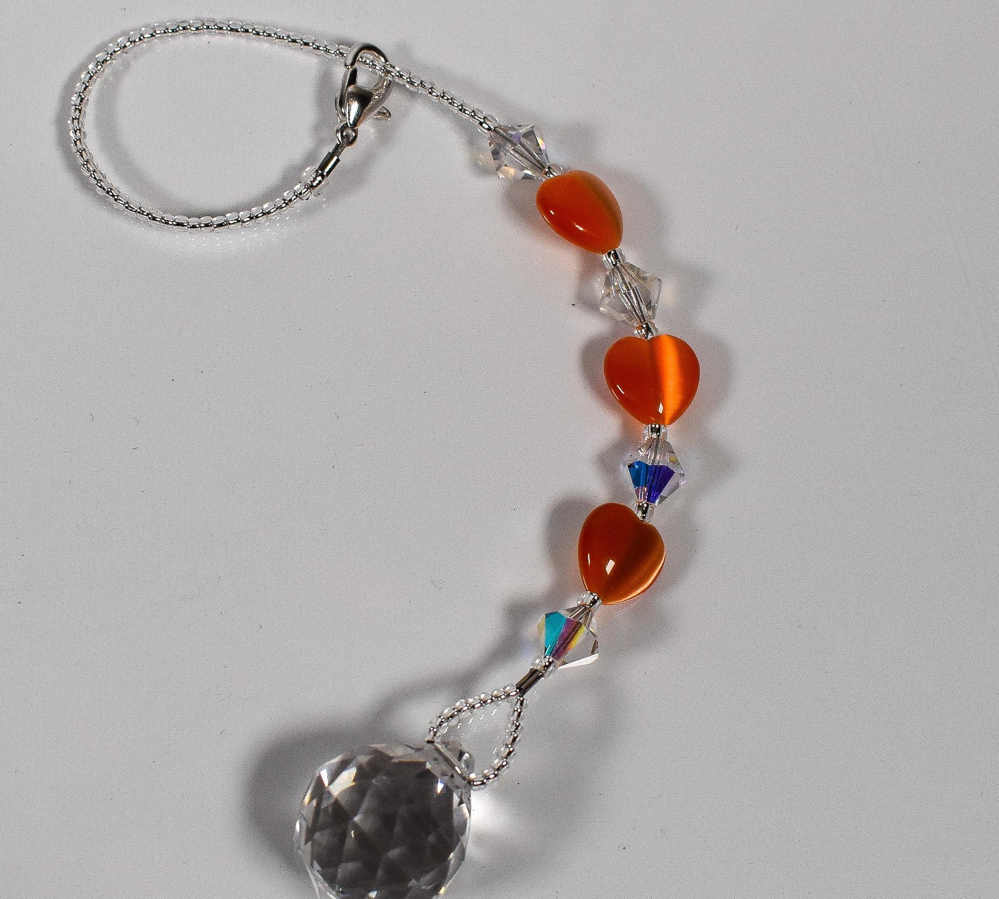 Small glass sun catcher made with small orange hearts, crystal bicones and a crystal prism, coiled up on white background.