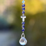 Crystal cross bead from Austria flanked by six Murano Glass beads in purple with silver foil, hanging vertically against blurred background, anchored by round glass crystal prism.