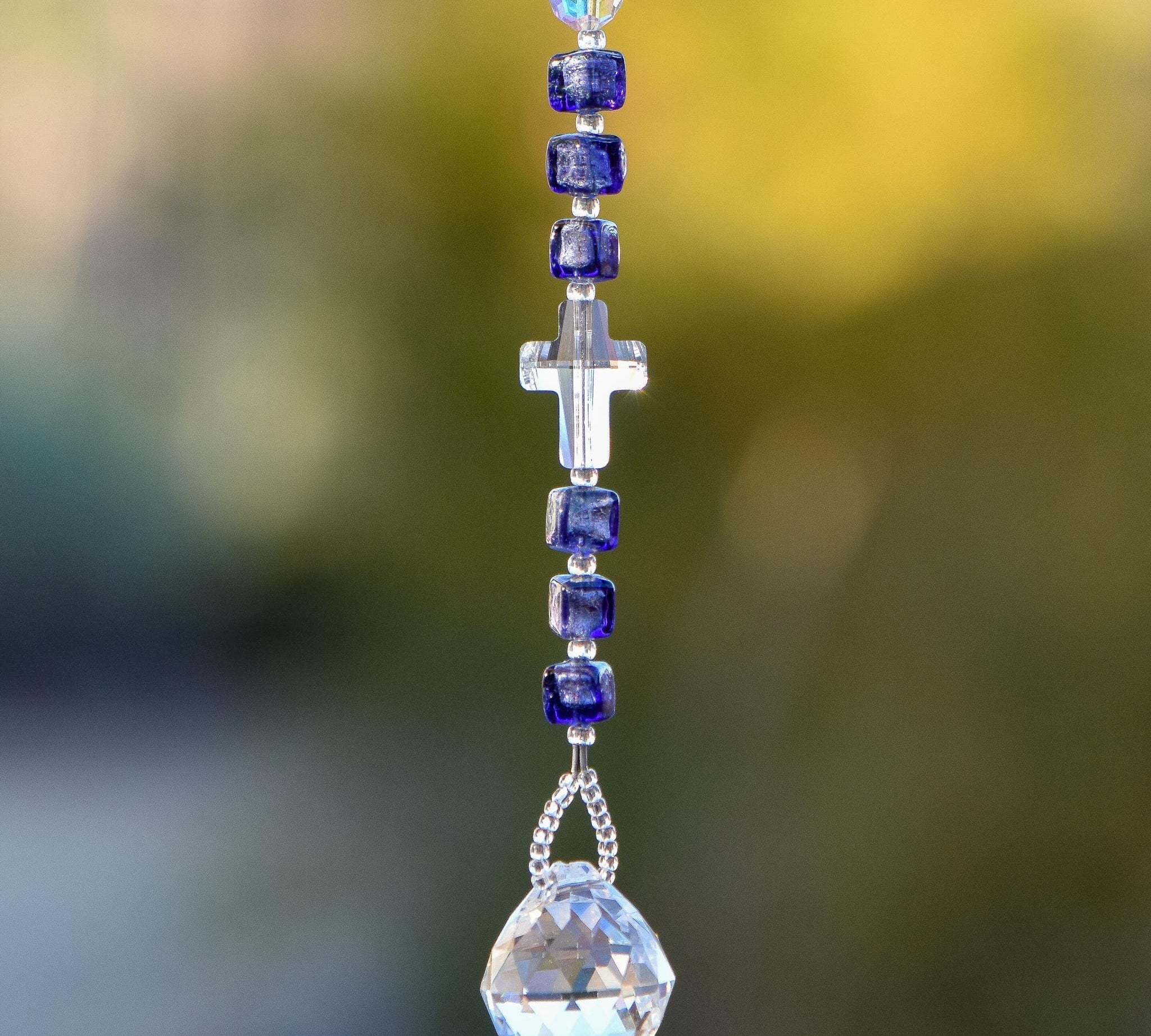 Crystal cross bead from Austria flanked by six Murano Glass beads in purple with silver foil, hanging vertically against blurred background, anchored by round glass crystal prism.