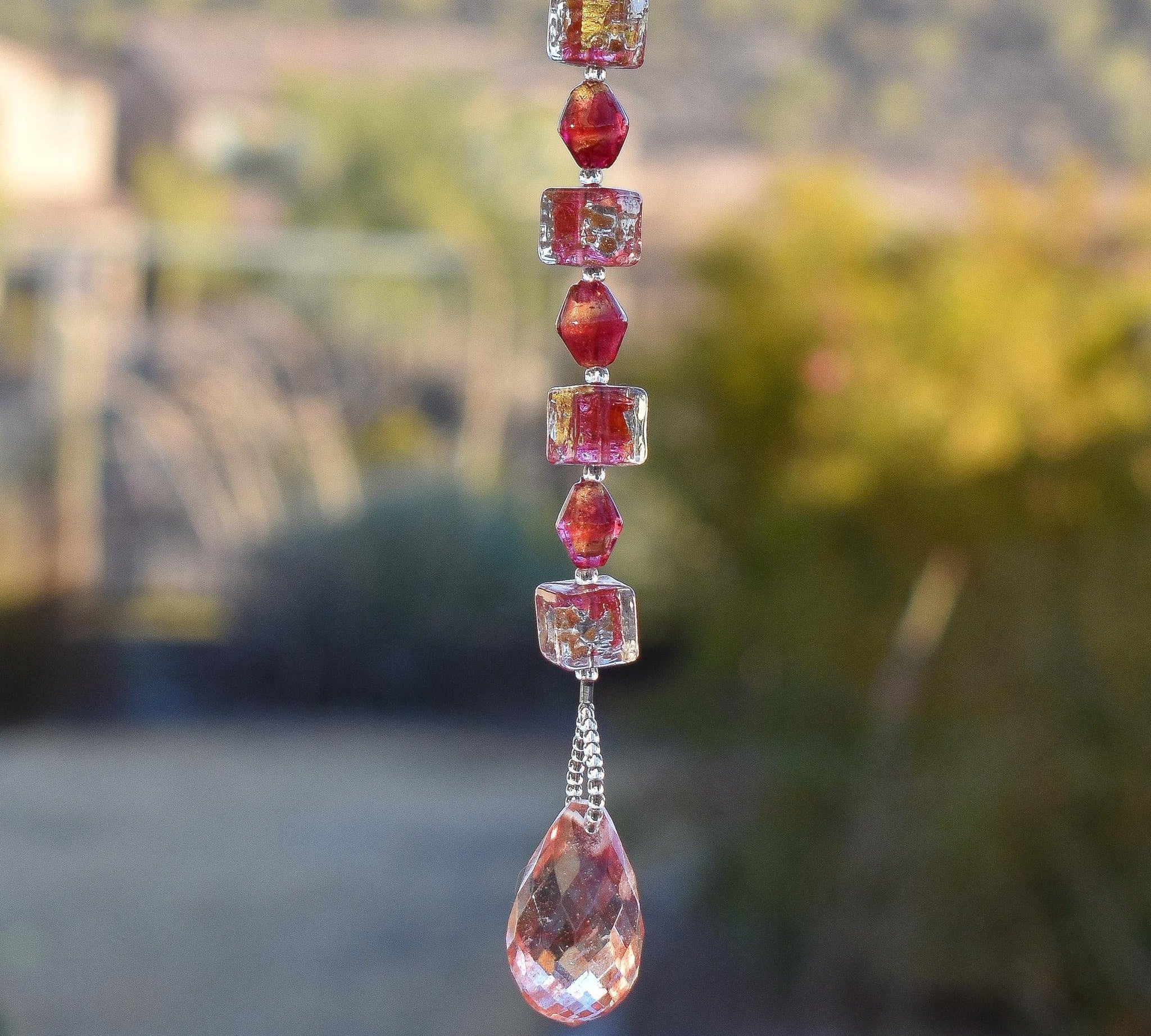 Small sun catcher hanging in landscape setting with 7 rubino pink murano glass beads and a large, pink, stone pendant