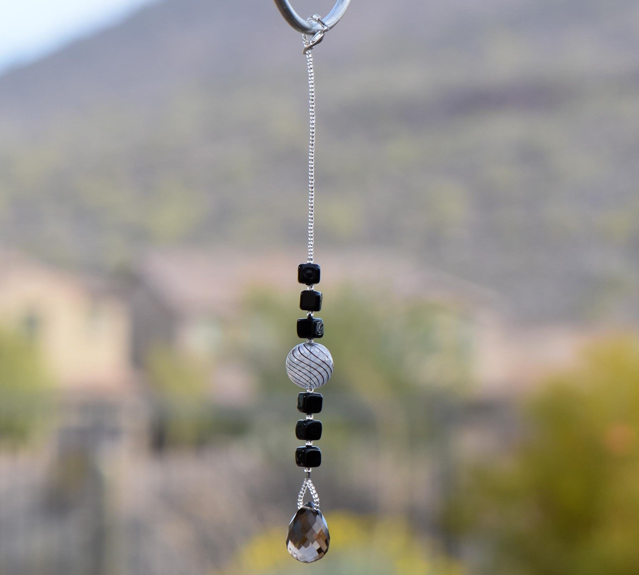 Small sun catcher hanging vertically in front of blurred background, made with six Murano glass cube beads flanking a large, round swirly bead and anchored by a large smoky quartz pendant