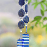 Hanging wind chime made with large thick lapis lazuli stone discs strung with glass beads, hanging vertically, anchored by two pieces of kiln formed clear glass with blue stripes.