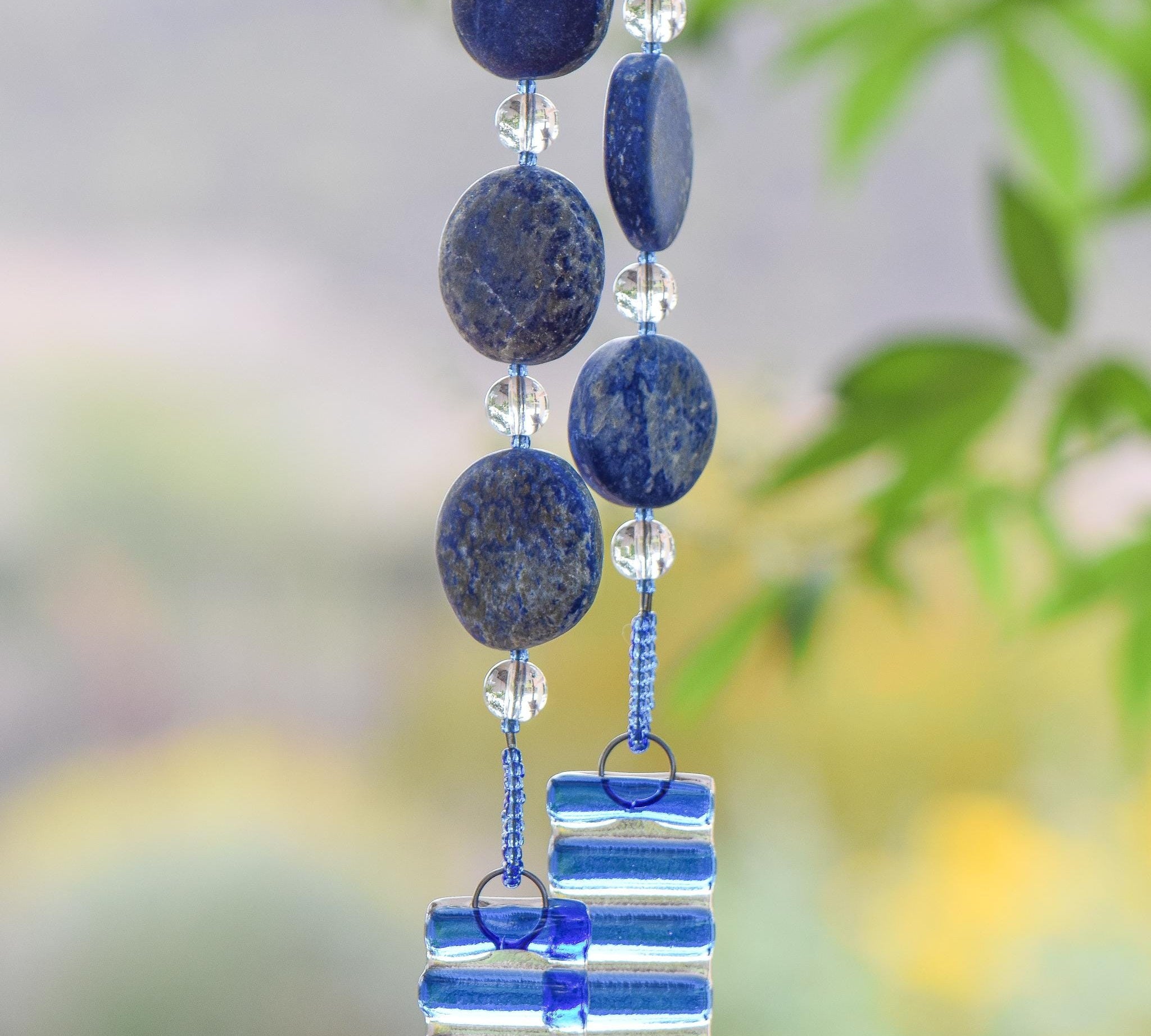 Hanging wind chime made with large thick lapis lazuli stone discs strung with glass beads, hanging vertically, anchored by two pieces of kiln formed clear glass with blue stripes.