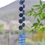 Hanging wind chime made with large thick lapis lazuli stone discs strung with glass beads, hanging vertically, anchored by two pieces of kiln formed clear glass with blue stripes.