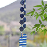 Hanging wind chime made with large thick lapis lazuli stone discs strung with glass beads, hanging vertically, anchored by two pieces of kiln formed clear glass with blue stripes.